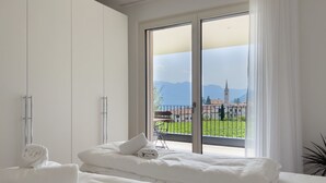 A hotel room with a large window, a bed with white linens, and a view of a town and mountains.