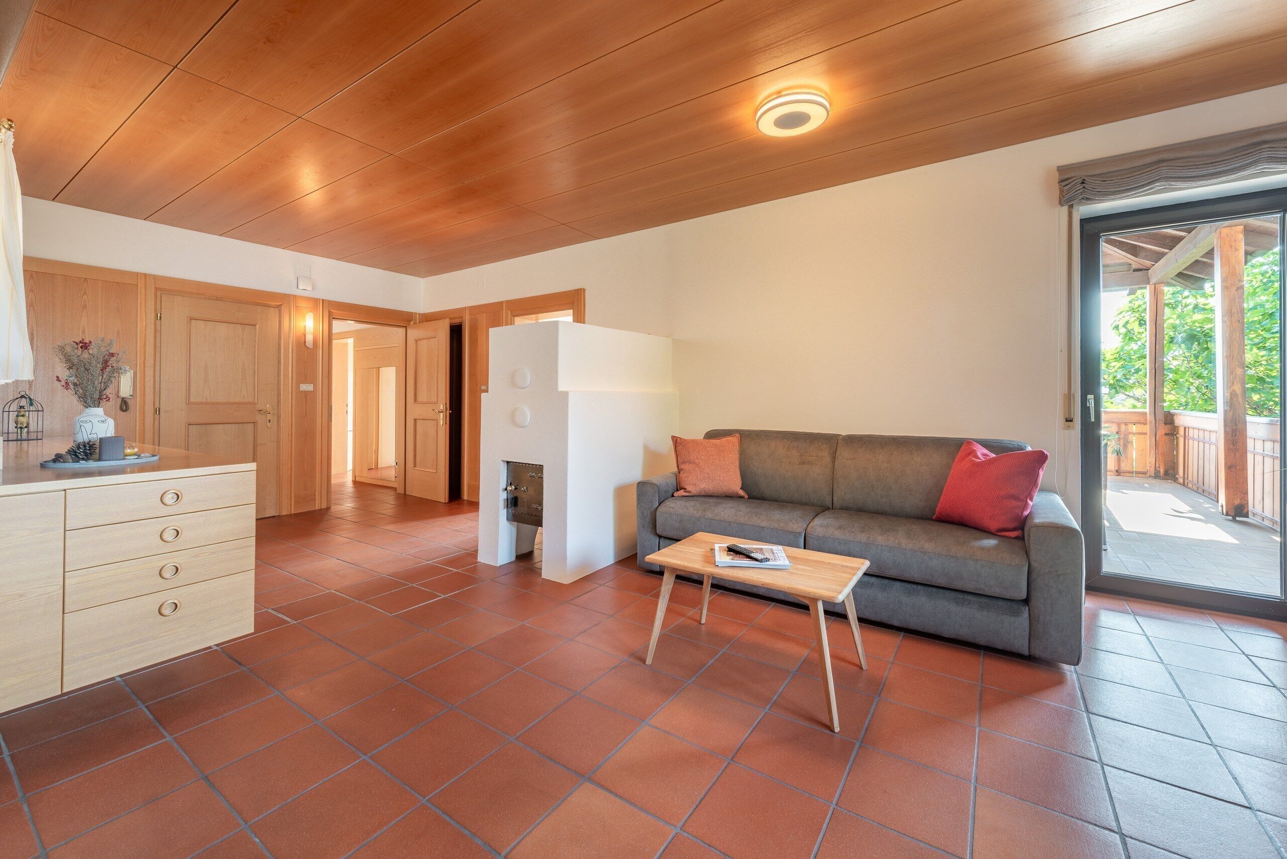 Un espace de vie moderne avec un plafond en bois, un canapé gris agrémenté de coussins rouges, une petite table basse en bois et un meuble blanc avec des tiroirs.