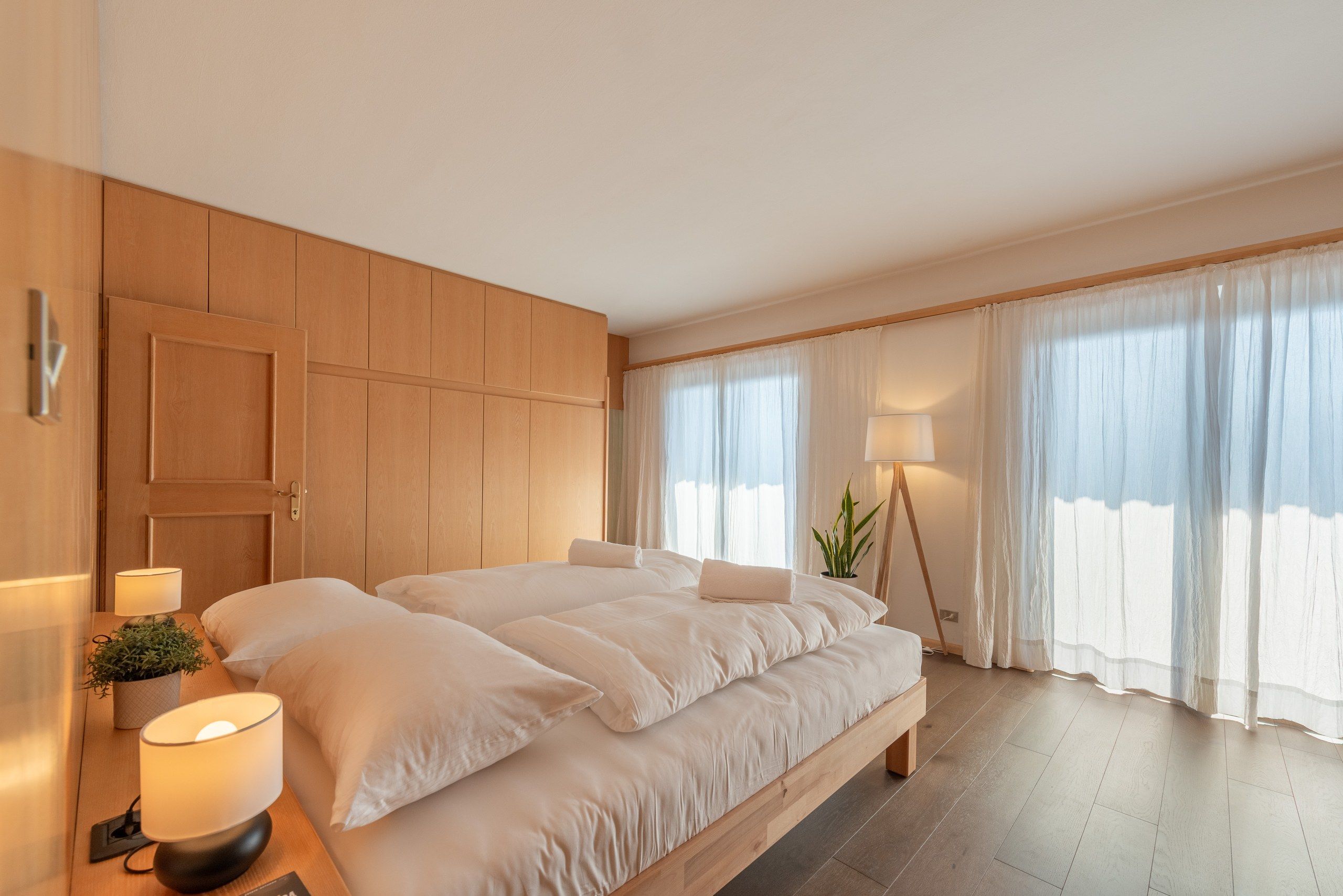 A hotel room with a large bed, bedside lamps, a potted plant, and a view through sheer curtains.
