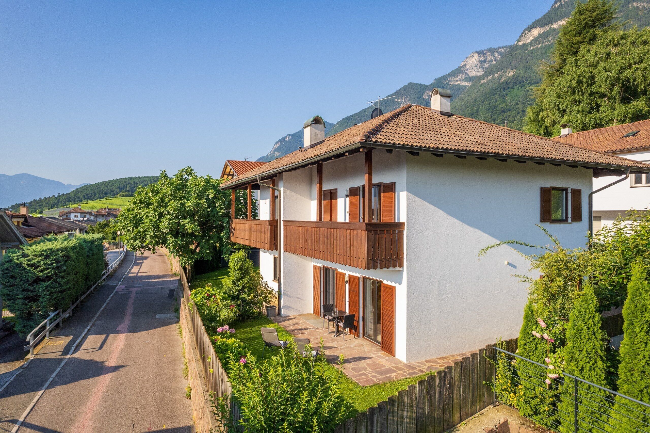 Une maison à deux étages avec un balcon, une allée pavée et une vue sur les montagnes en arrière-plan.