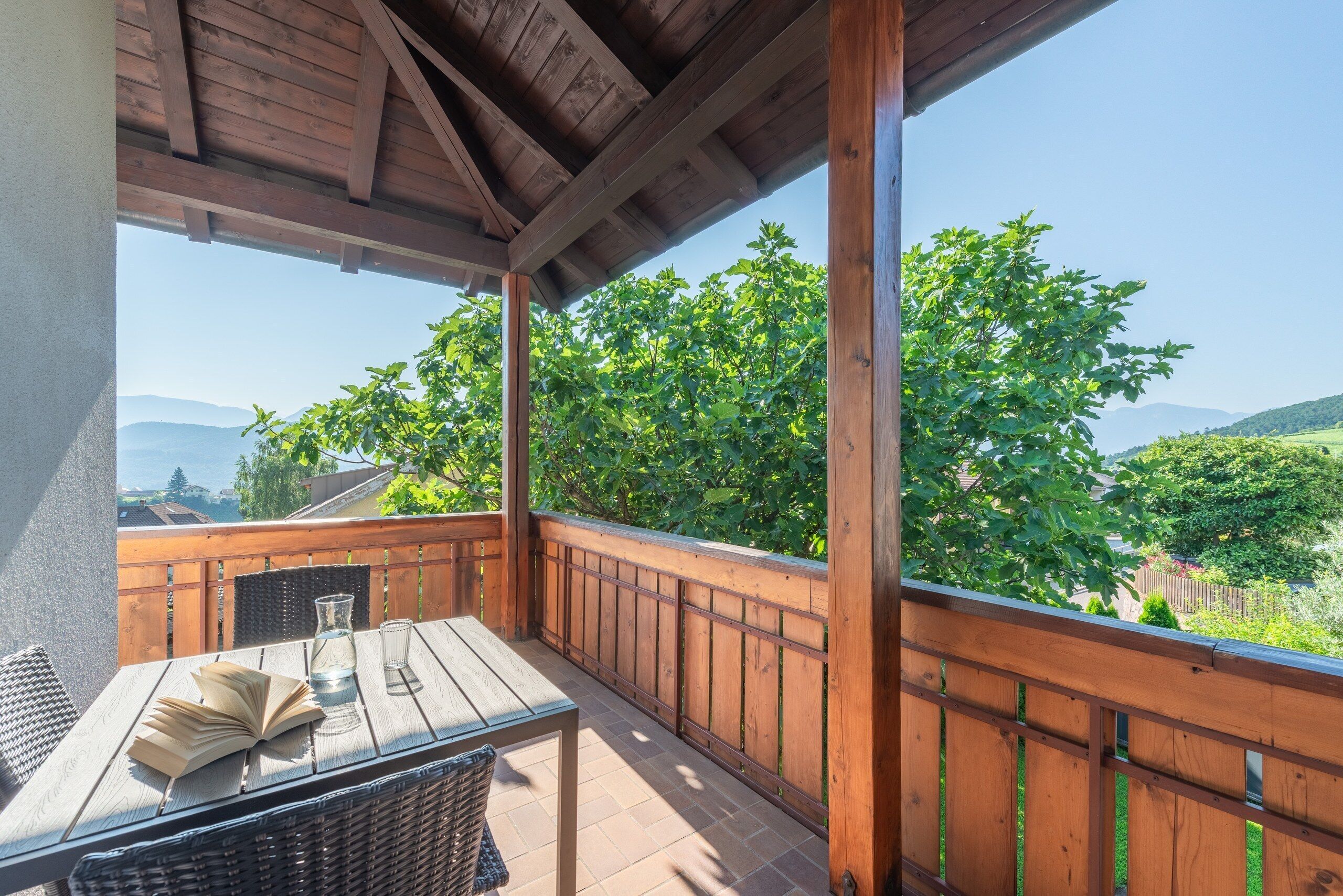 A wooden balcony with a table and chairs, overlooking a lush green landscape and distant mountains.