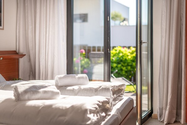 A hotel room with a bed, a wooden headboard, a window with curtains, and a view of greenery outside.