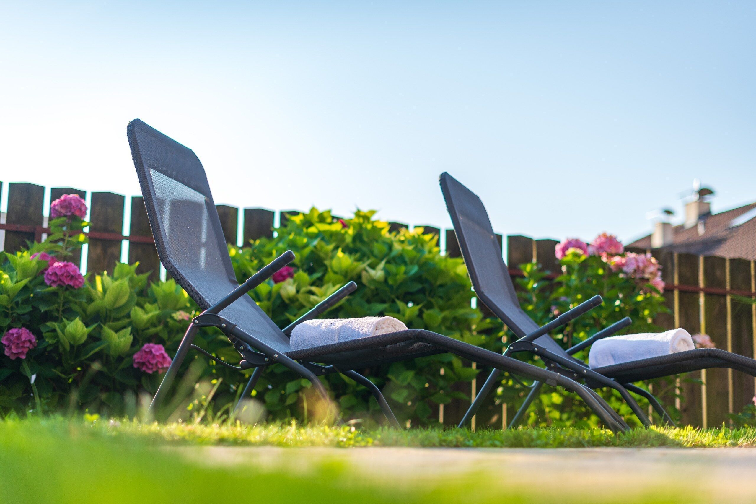 Twee loungestoelen met handdoeken, geplaatst in een tuin met een houten omheining en bloeiende planten op de achtergrond.