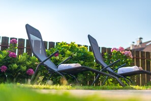 Dos sillones de descanso con toallas, ubicados en un jardín con un cerco de madera y plantas florecientes al fondo.