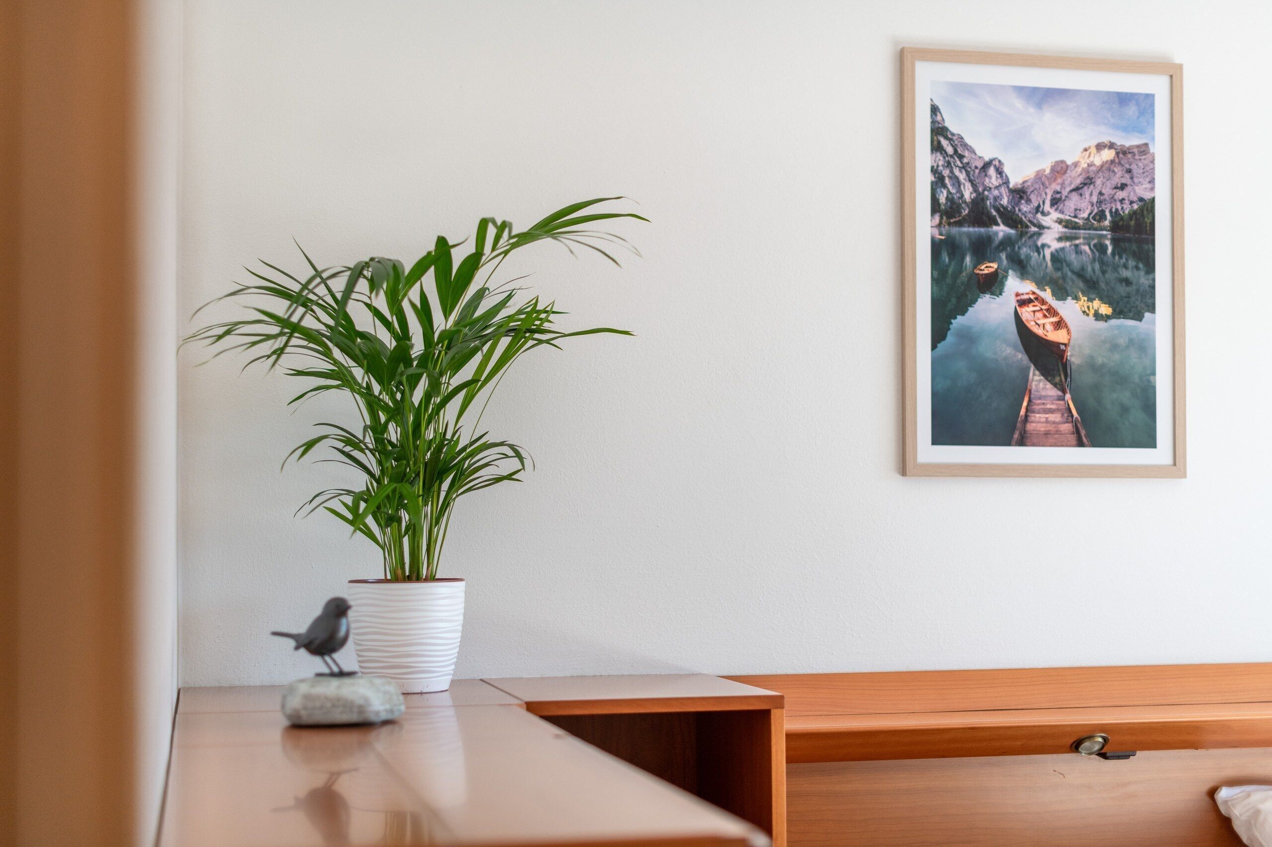Una habitación con un escritorio de madera, un jarrón blanco con una planta, un cuadro con un paisaje de un lago y una pequeña figurita de pájaro.