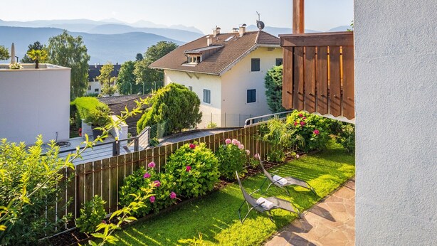 Ein Wohnhaus mit einem gepflegten Garten, einer Terrasse mit Liegestuhl und Blick auf ferne Berge.