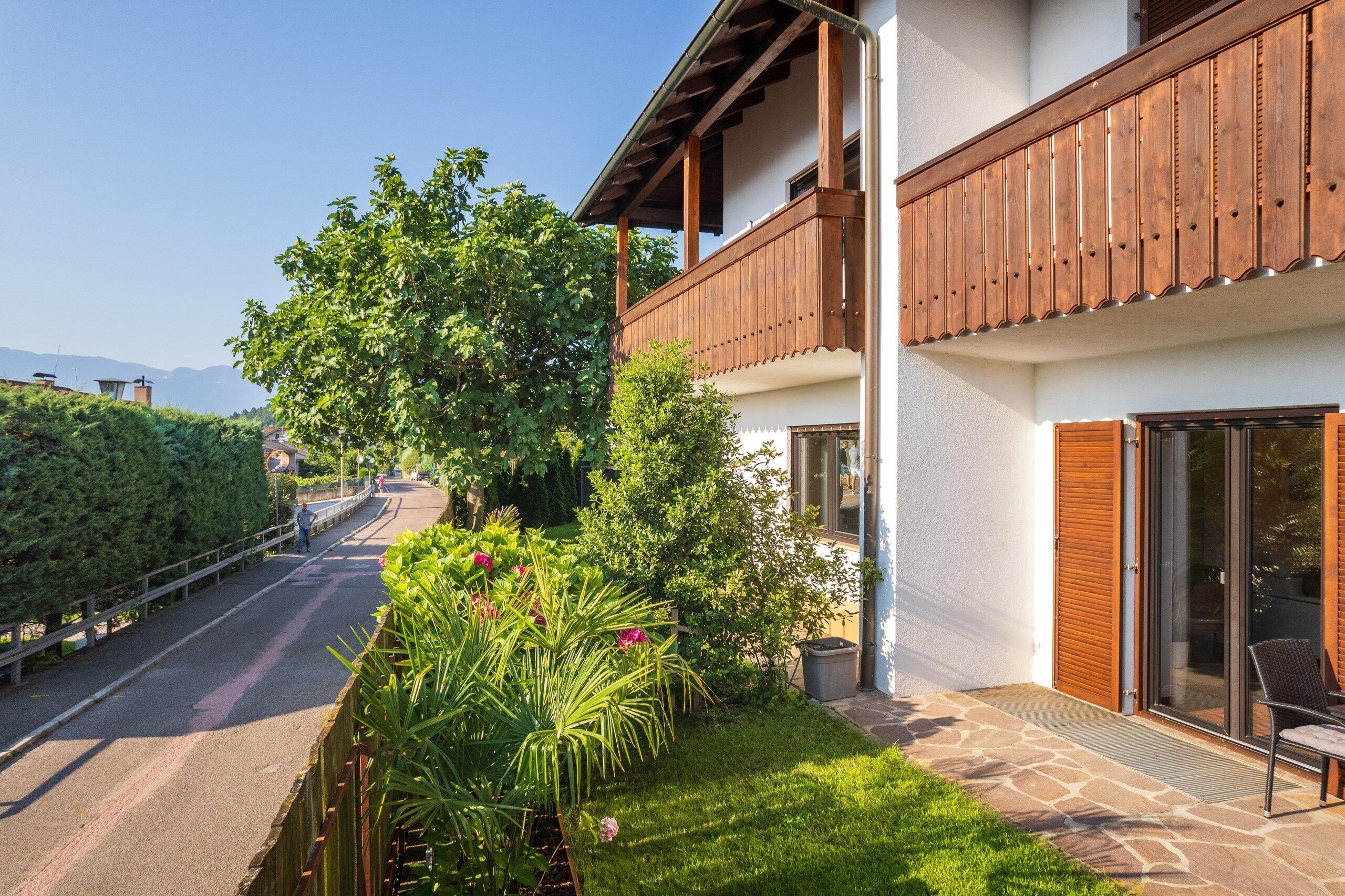 Un edificio a due piani con un balcone, un giardino con diverse piante e una strada sullo sfondo.