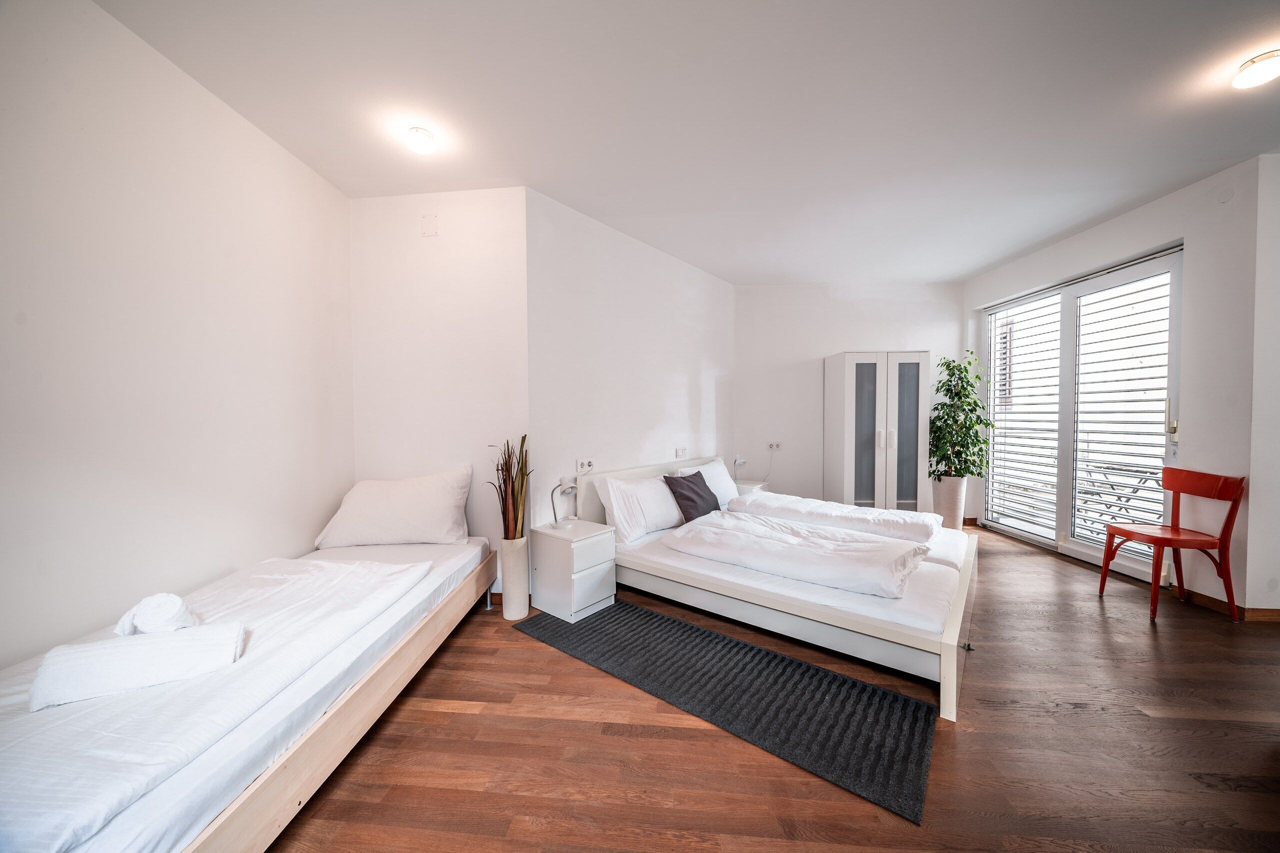 Een moderne slaapkamer met een bed, nachtkastjes, een stoel en een groot raam met jaloezieën.