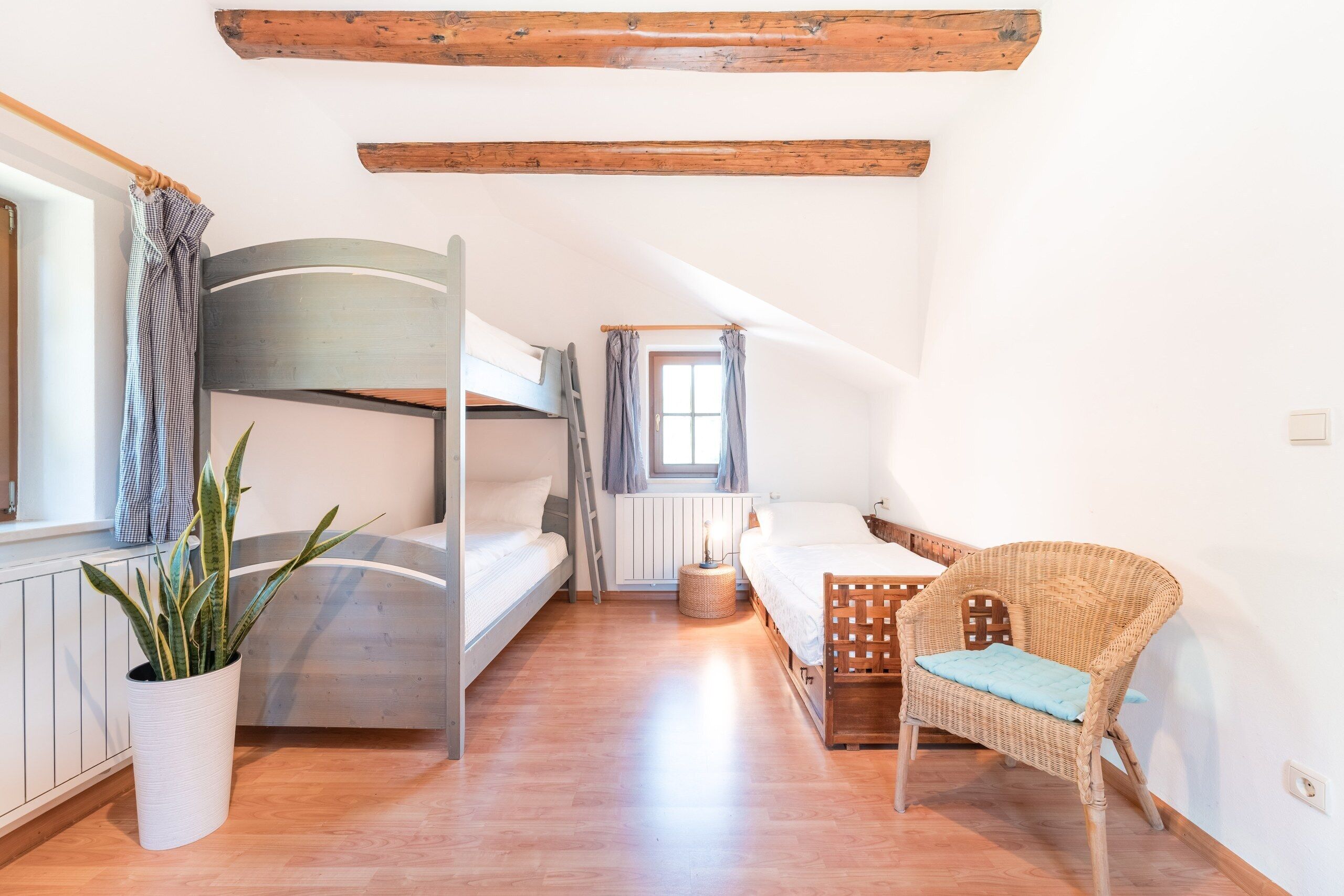 Une chambre avec un lit superposé, une chaise en osier, une plante en pot et une fenêtre avec des rideaux.