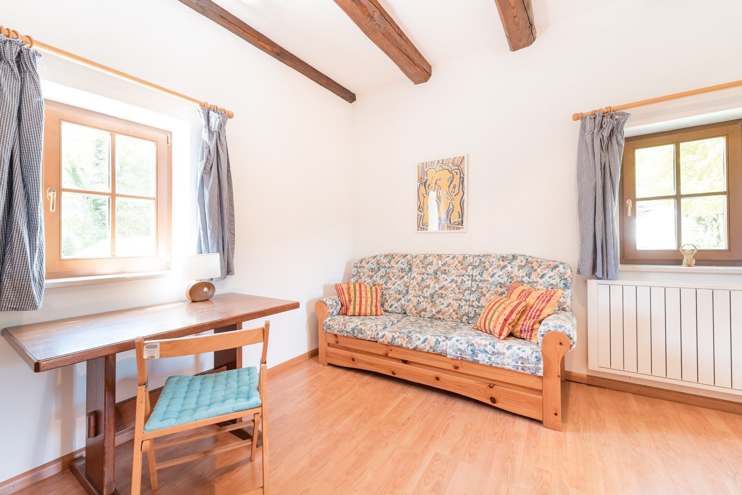 Una habitación con piso de madera, un sofá con tapicería floral, una mesa de madera y una silla con cojín azul.