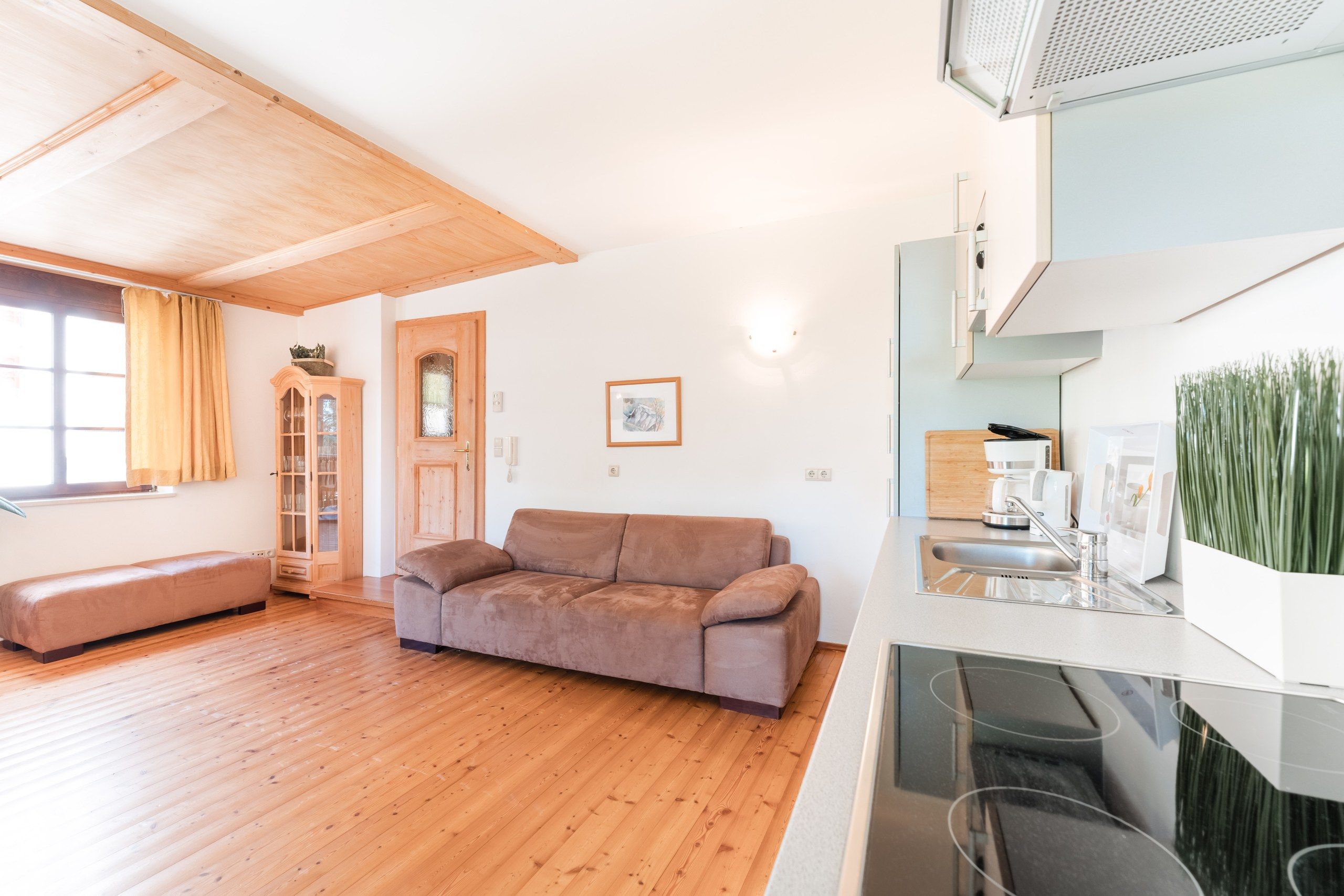Un salon moderne avec un plancher en bois, un canapé marron, un meuble en bois et un coin cuisine équipé d’un évier et d’une machine à café.