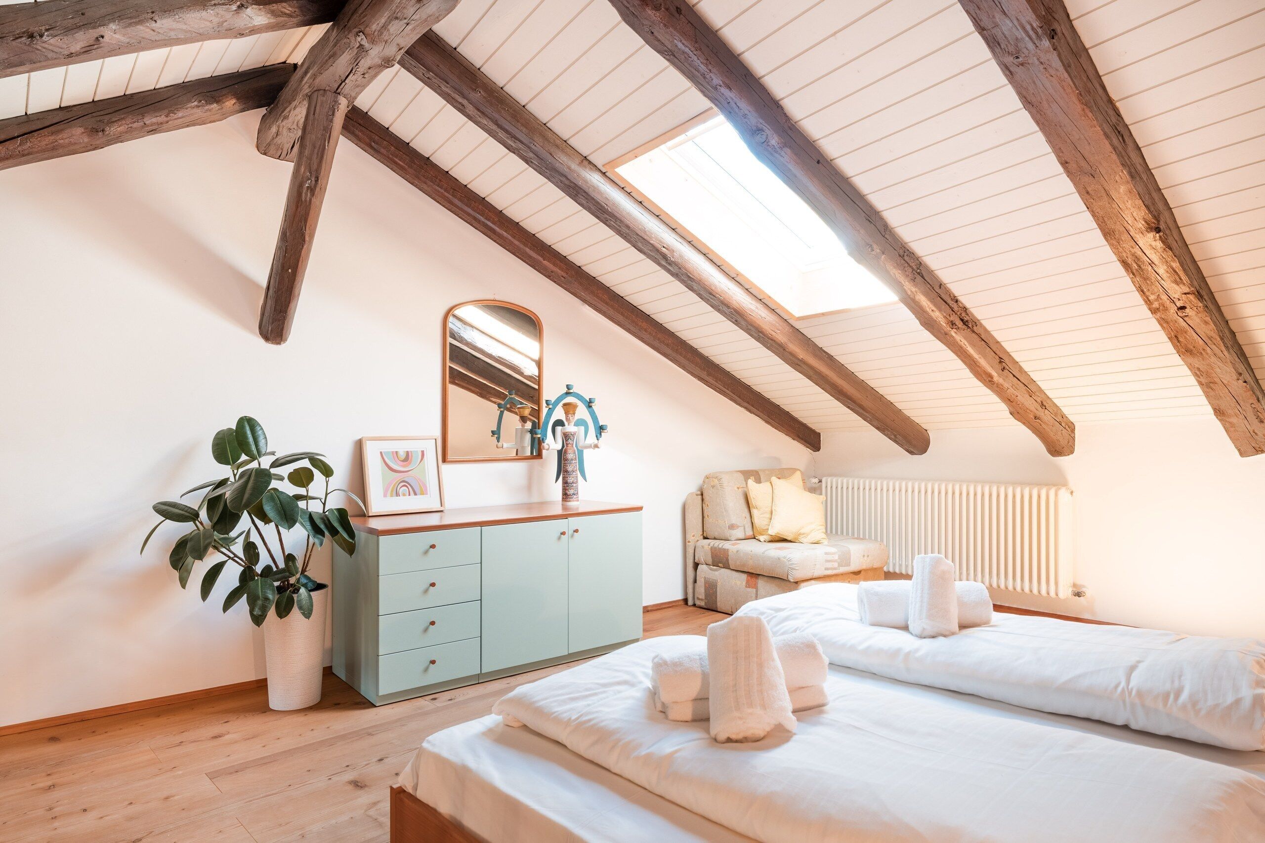 Une chambre avec un plafond en pente, des poutres en bois, une commode, un miroir, une plante, un canapé et un lit avec des serviettes.
