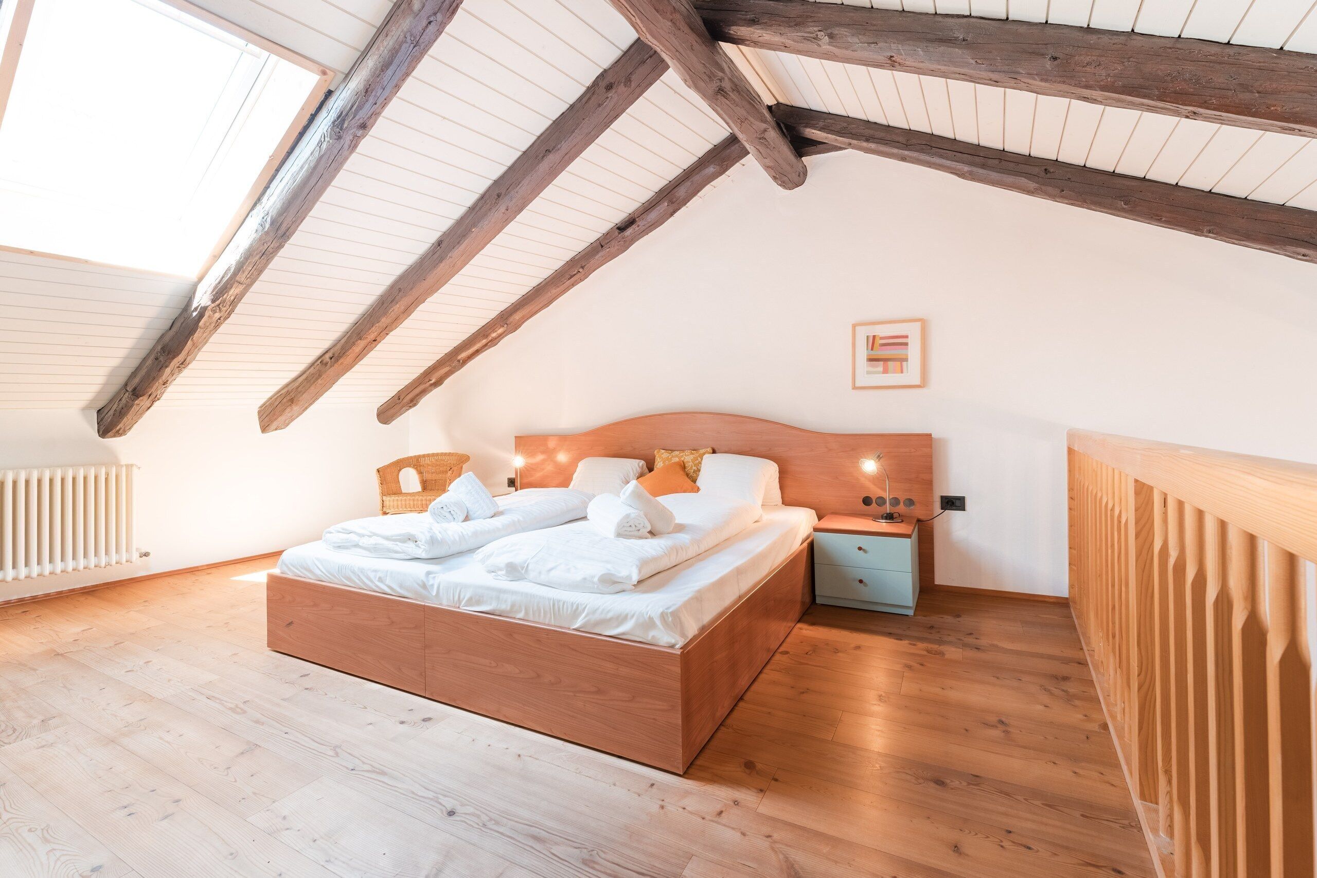 Une chambre avec un plafond en pente, des poutres en bois, un lit avec du linge de maison blanc, une table de chevet et un tableau encadré au mur.