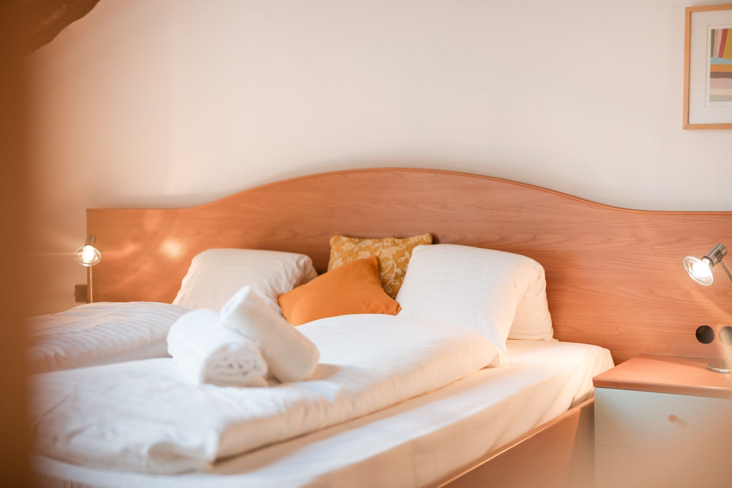A neatly made bed with white linens and a white towel, a wooden headboard, and two bedside lamps.