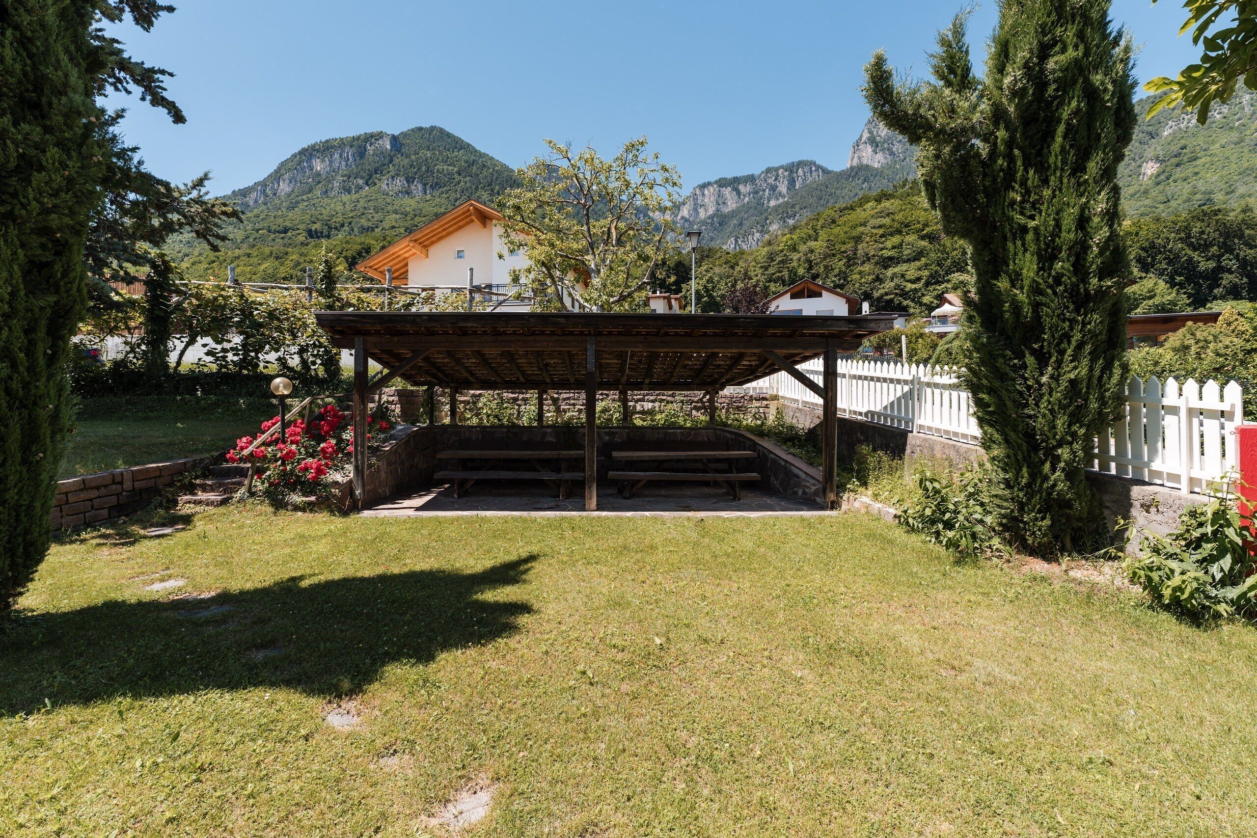 A covered outdoor seating area with a wooden structure, surrounded by grass and trees, with mountains in the background.