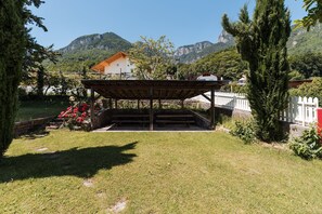 Un área de estar al aire libre cubierta con una estructura de madera, rodeada de césped y árboles, con montañas al fondo.