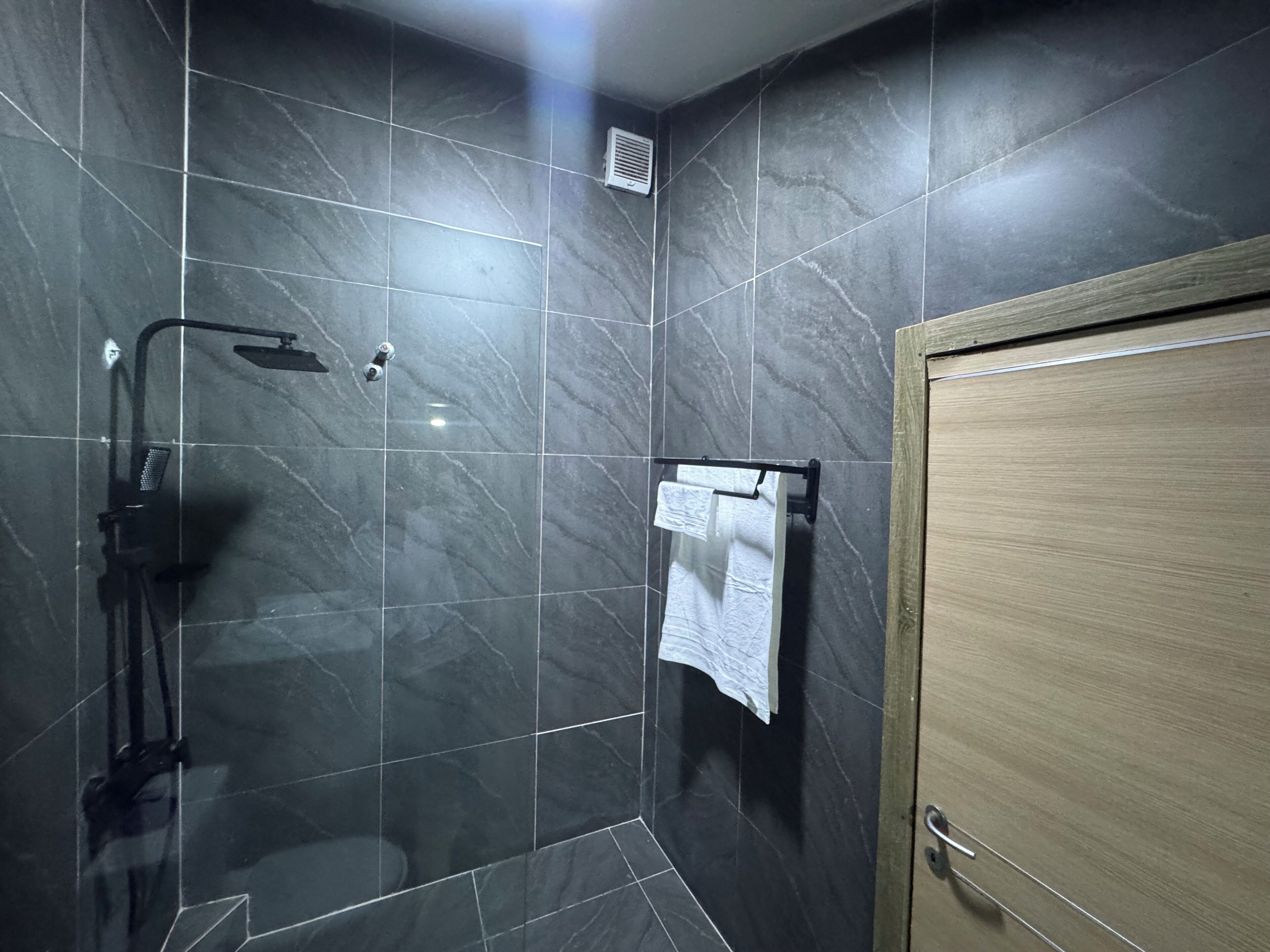 Un baño moderno con baldosas de mármol oscuro, ducha con mampara de vidrio, barra para toallas y puerta de madera.