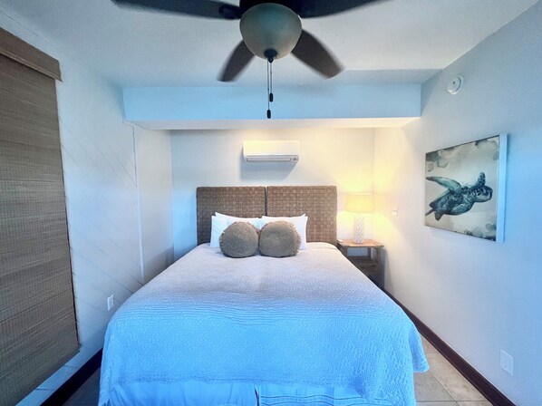 A hotel room with a bed, a ceiling fan, an air conditioning unit, a nightstand with a lamp, and a wall-mounted artwork.