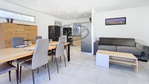 A modern living room with a grey sofa, a wooden dining table with chairs, and a kitchen area with cabinets and a refrigerator. - Modern house for 4 people close to Cavalière beach in Le Lavandou (Le Lavandou)