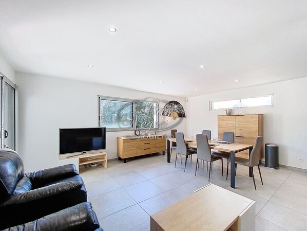 A modern living room with a flat-screen TV, a leather sofa, a dining area with a wooden table and chairs, and a kitchen area with cabinets and a window.