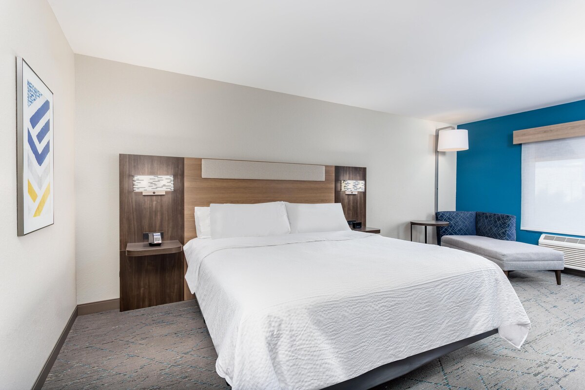 Quarto de hotel com uma cama grande, mesas de cabeceira, escrivaninha, uma cadeira e janela com persianas.