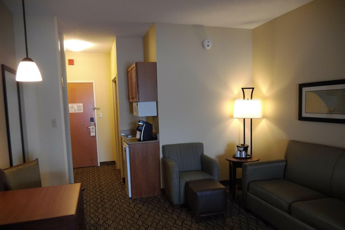 Habitación de hotel con kitchenette, una mesa de comedor, un sofá, un pequeño reposapiés, una lámpara y un cuadro en la pared.