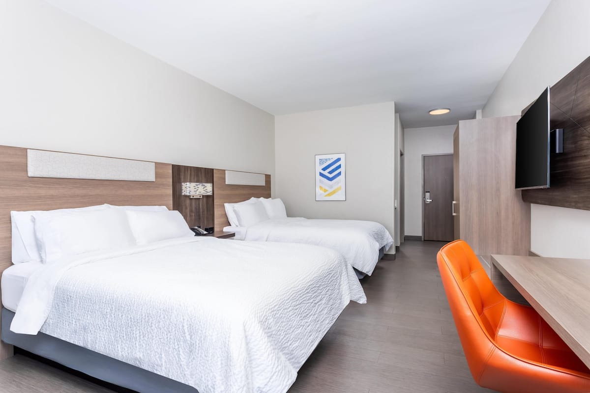 A hotel room with two beds, a TV, a wooden headboard, and a wooden dining table with an orange chair.