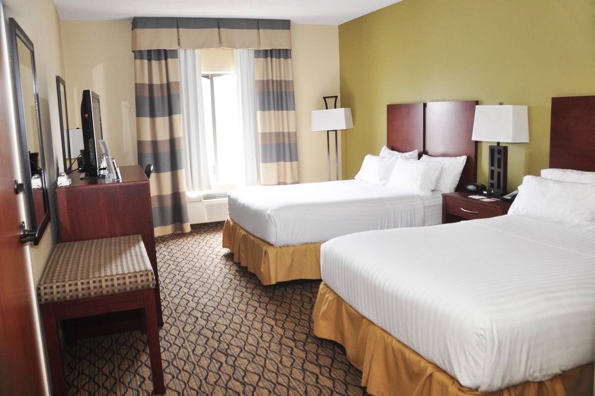A hotel room with two beds, a desk with a chair, a desk lamp, a bedside table with a lamp, and a window with curtains.