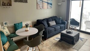 A living room with a blue sofa, a round table, and a coffee table. There is a painting of a beach scene on the wall, and a sign that says "cause down the shore everything's alright."
