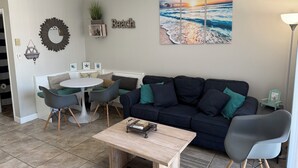 A living room with a round table, a dark sofa, a coffee table, and a wall-mounted artwork of a beach scene.