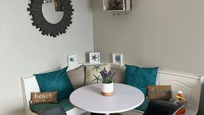 A cozy living room with a white sofa, two black chairs, a round white table, and a small potted plant. A decorative mirror and a wall-mounted shelf with a plant and starfish are also visible.