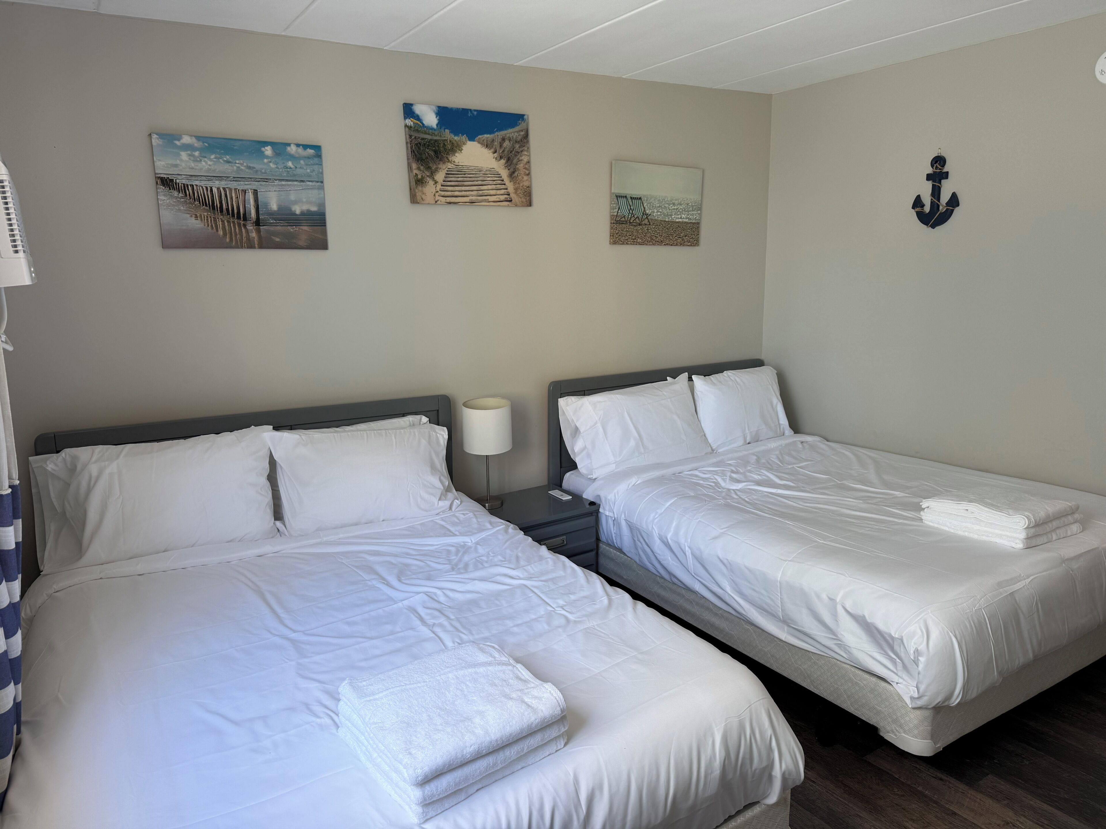 A hotel room with two beds, a nightstand with a lamp, and wall art depicting beach scenes.