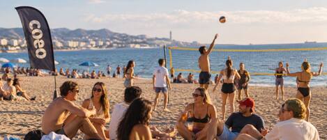 Escena de playa con gente sentada y jugando voleibol, una bandera y un paisaje urbano al fondo.