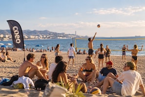 Eine Strandansicht mit Personen, die sitzen und Volleyball spielen, einer Flagge und einer Stadtkulisse im Hintergrund.