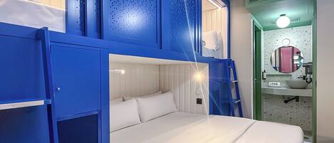 Habitación con literas, una cama blanca, un ventanal pequeño y un baño con espejo y lavabo.