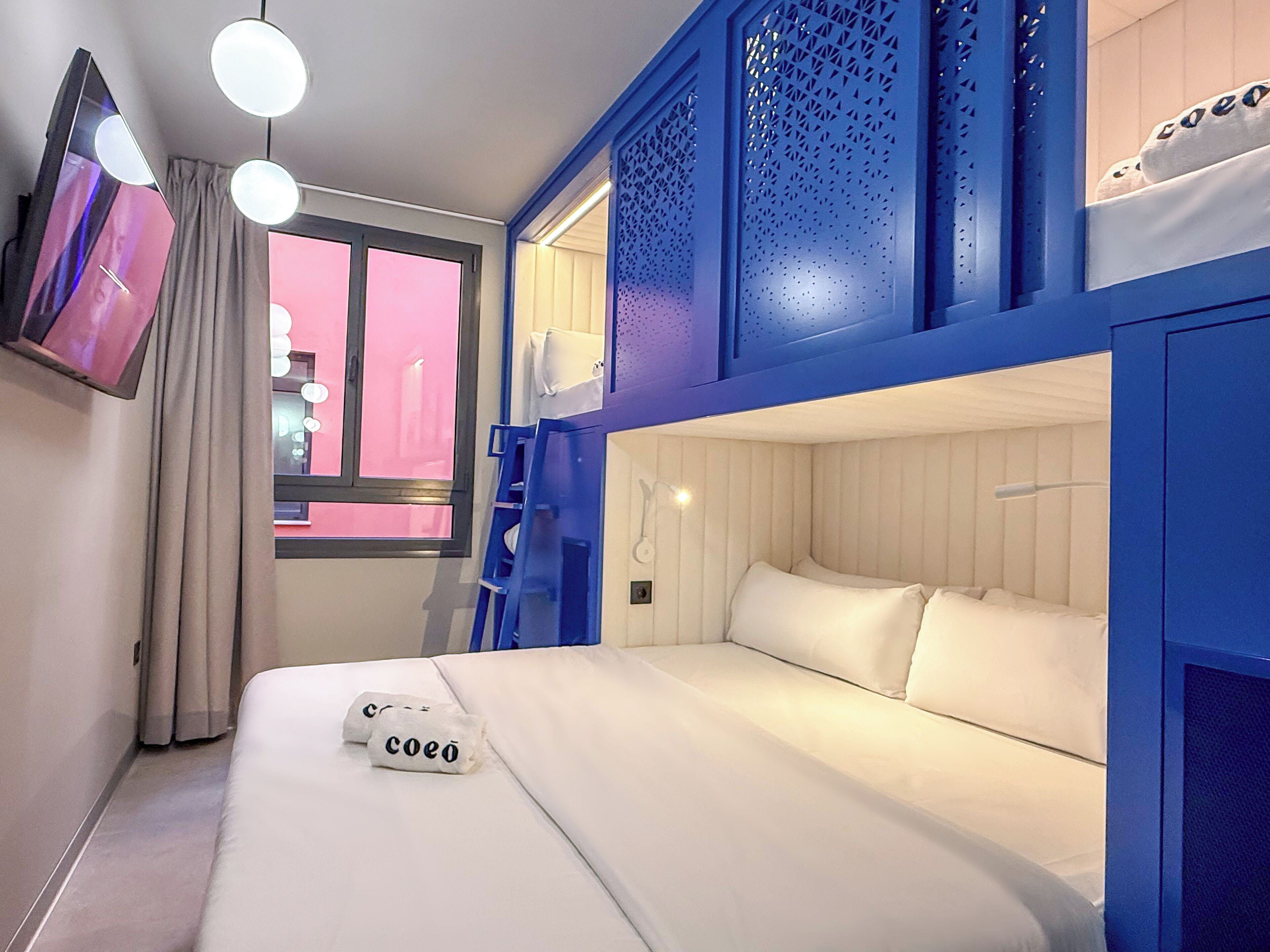 A hotel room with a bunk bed, a window with curtains, a TV mounted on the wall, and a bedside table with towels.