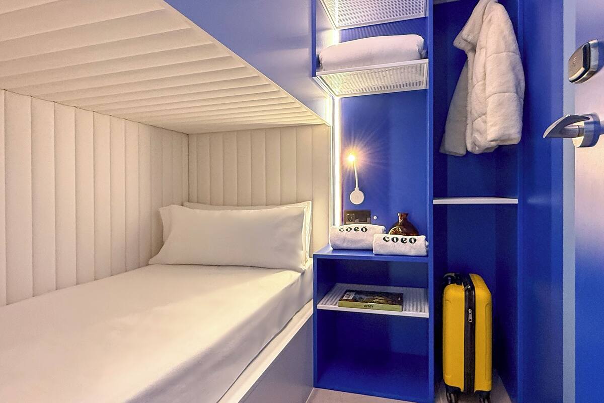 Een kleine hotelkamer met een enkel bed, een klein plankje met handdoeken, een koffer en een wandlamp.