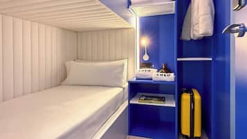 A compact hotel room with a single bed, a small shelf with towels, a suitcase, and a wall-mounted lamp.
