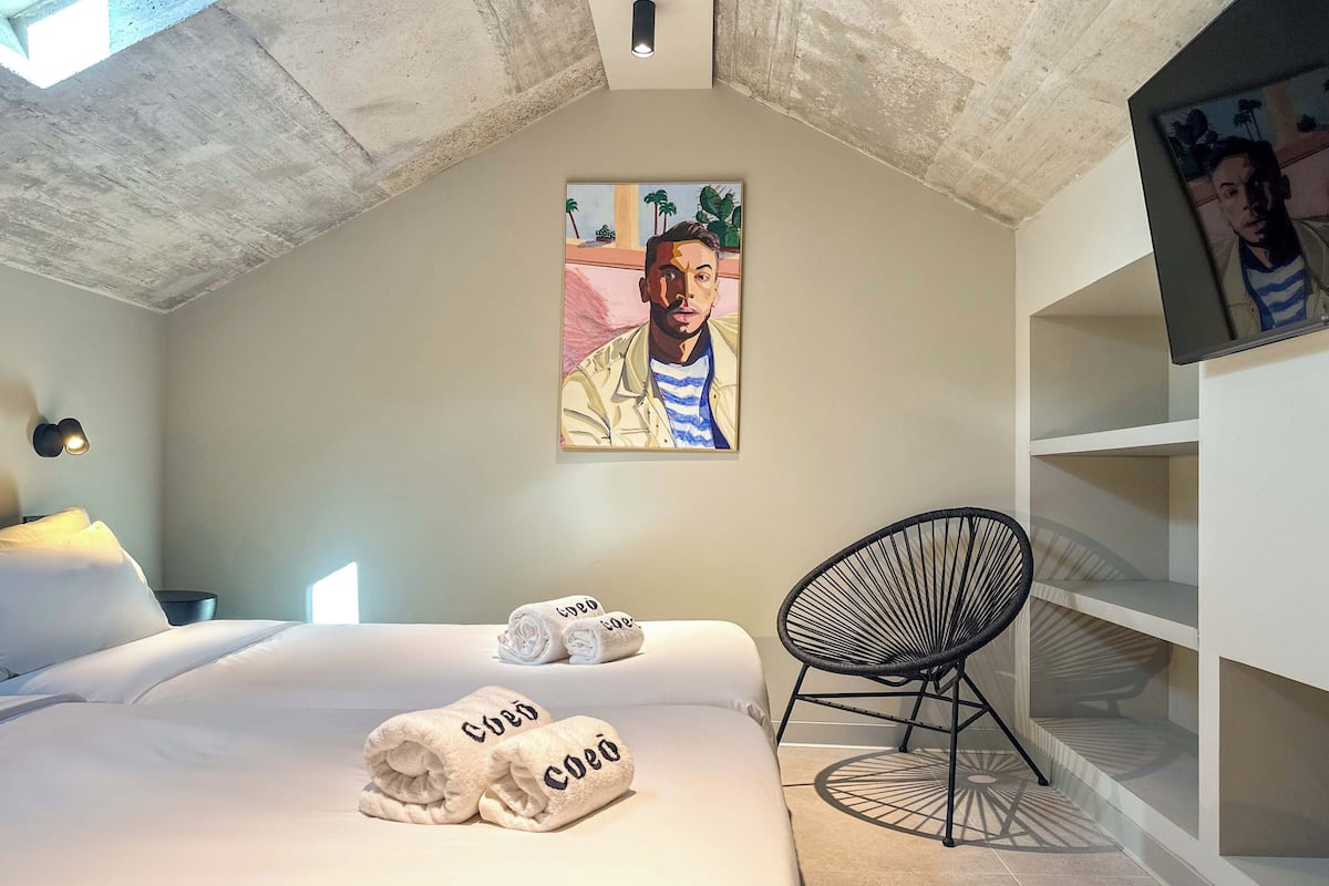 Een slaapkamer met een bed, twee opgerolde handdoeken, een stoel, een schilderij en een plank.