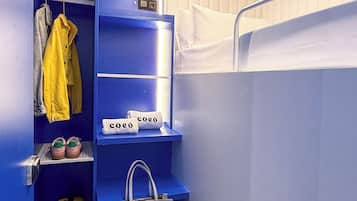 Una habitación pequeña con una cama litera, un armario con ropa colgada, un estante con una bolsa y una maleta.