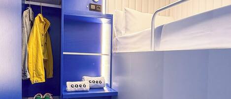 A compact room with a bunk bed, a wardrobe with hanging clothes, a shelf with a bag, and a suitcase.
