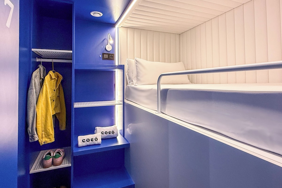 Een kleine hotelkamer met een stapelbed, een kledingkast met kleren, een plank met handdoeken en een klein plankje met schoenen.