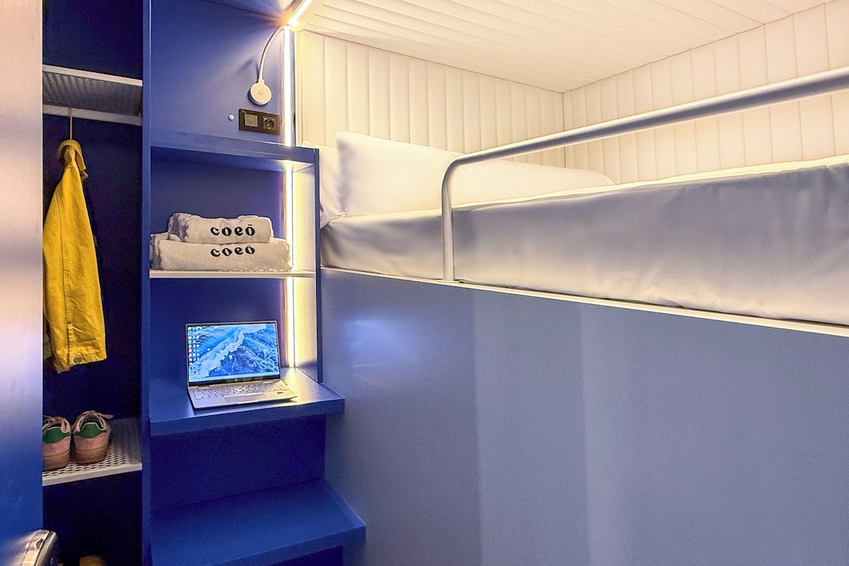 Een compacte, moderne kamer met een stapelbed, een klein bureau met een laptop en planken met persoonlijke spullen.