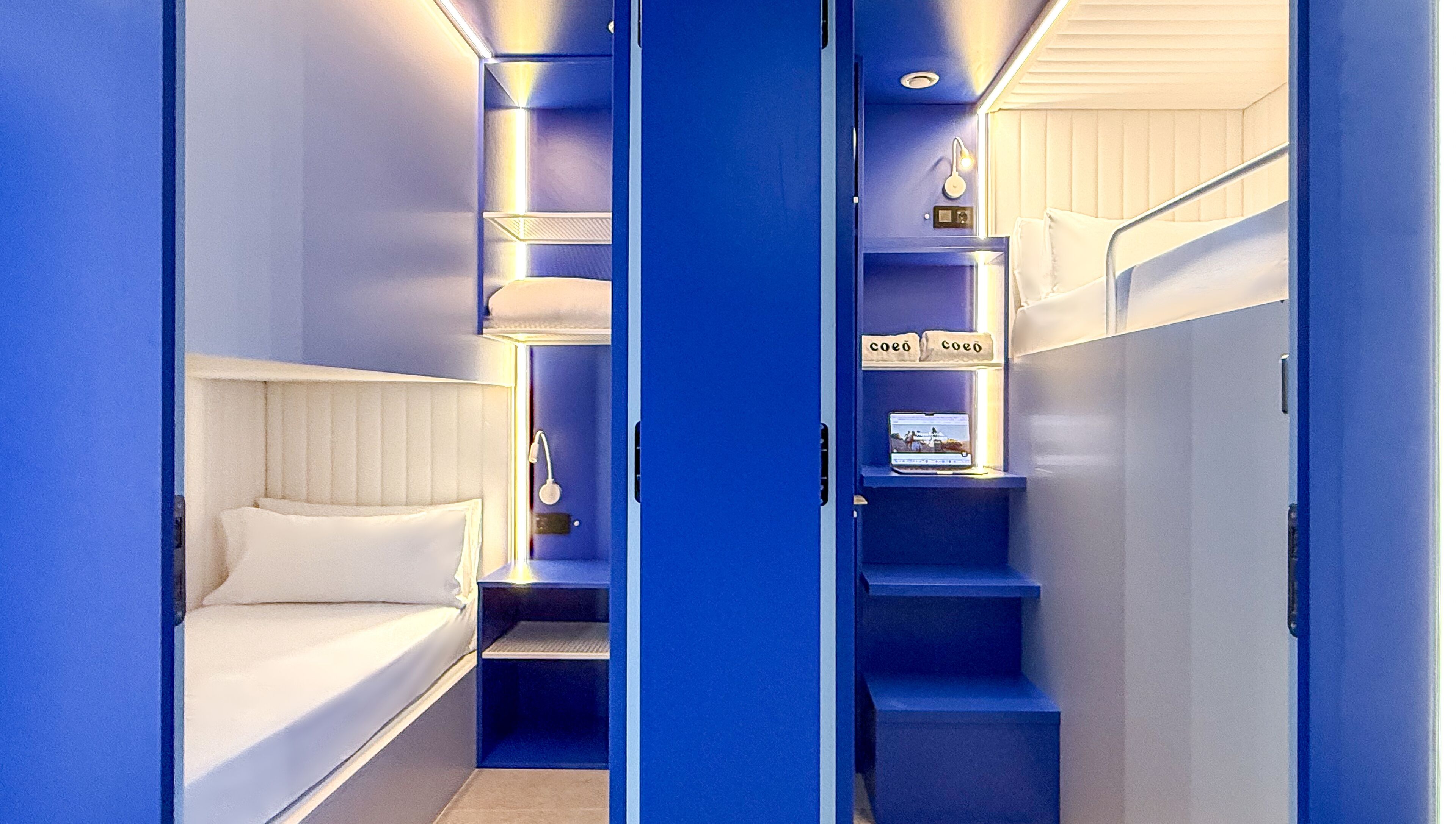 A compact hotel room with a bunk bed, a small desk with a laptop, and a shelf with decorative items.