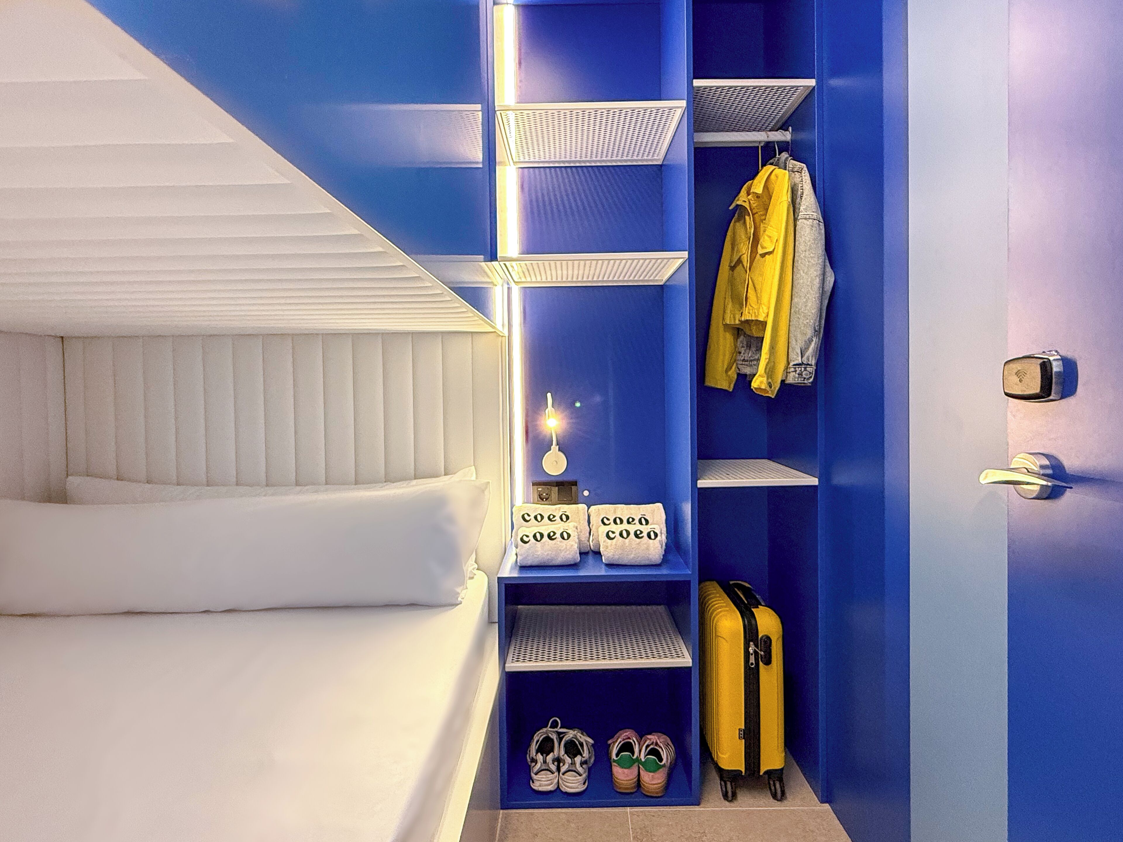 Una habitación de hotel con una cama, un armario con estantes, una maleta y un par de zapatos.