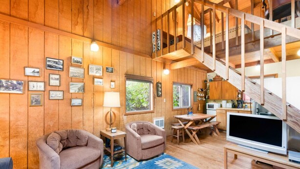 A cozy cabin interior with wooden walls, a staircase, a dining area, and a TV.