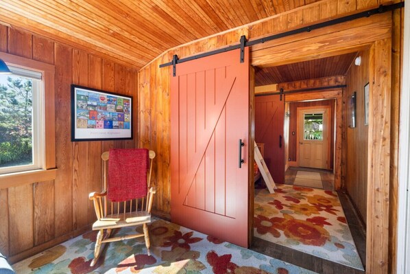 A wooden interior with a red sliding door, a framed picture on the wall, a wooden chair with a red towel, and a floral carpet.