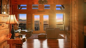 Interior de una cabaña de madera con chimenea, un sofá, una lámpara y vistas a las montañas a través de las ventanas.