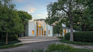 Una casa moderna con fachada de vidrio, rodeada por un jardín cuidado y un camino de entrada pavimentado.