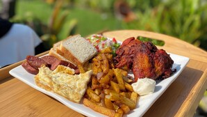 A plate of food including eggs, sausage, fries, and a piece of meat on a wooden table outdoors. - Emerald Villa Cafe & Restaurant (Kigali)